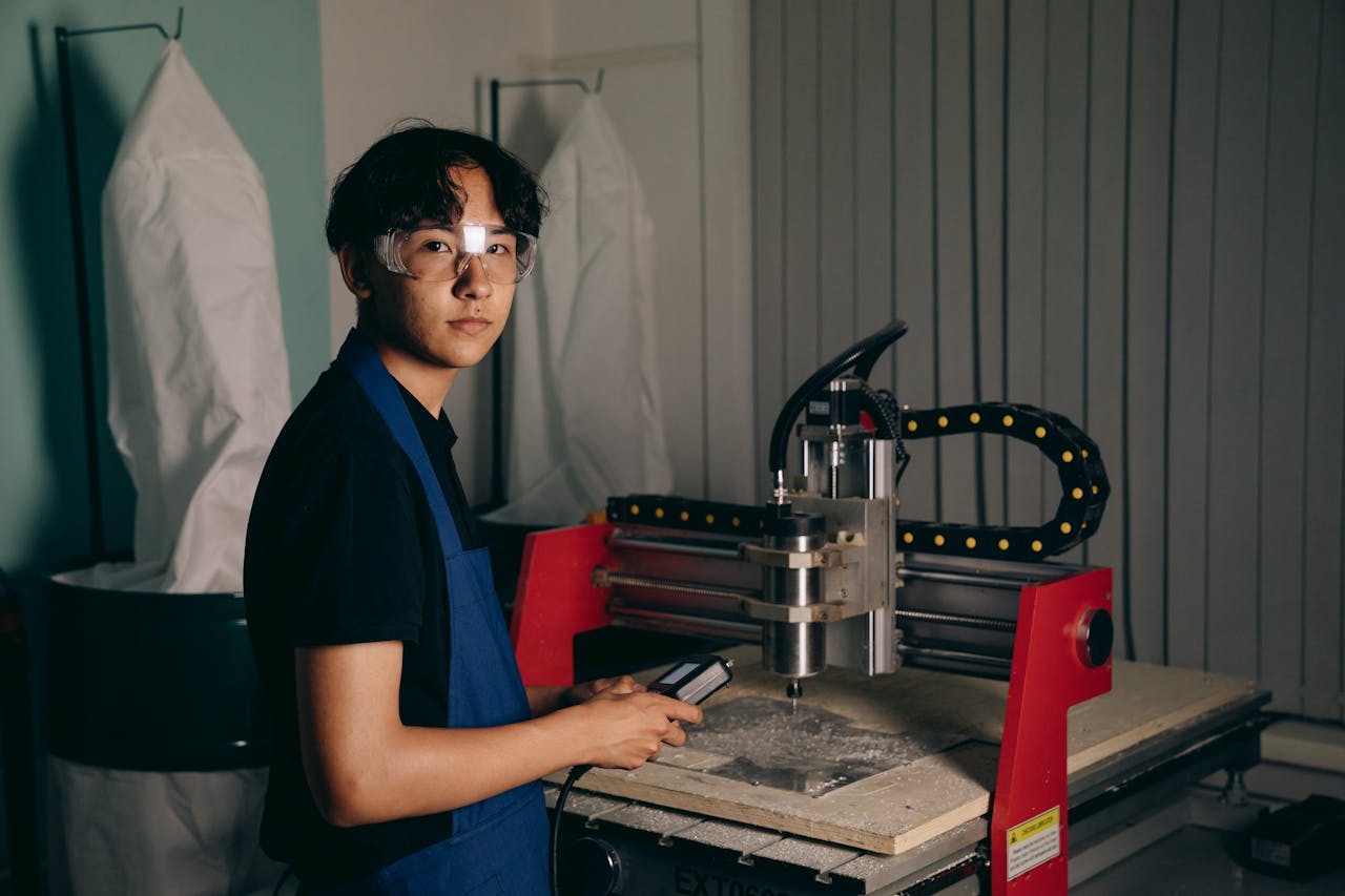 team-03 Portrait of a young worker operating a CNC machine in a workshop, wearing safety glasses and an apron.